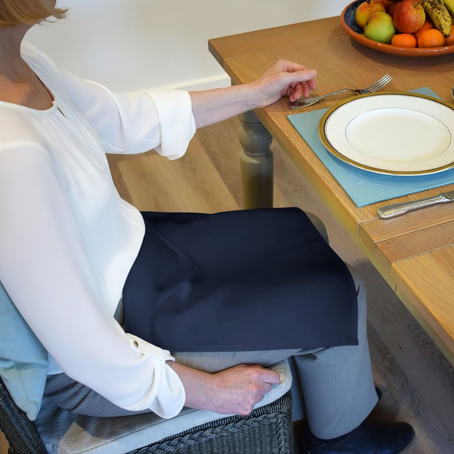 Lady sitting at a table with a plate and fork, wearing a white blouse and grey trousers with a Care Designs navywaterproof napkin on their lap.