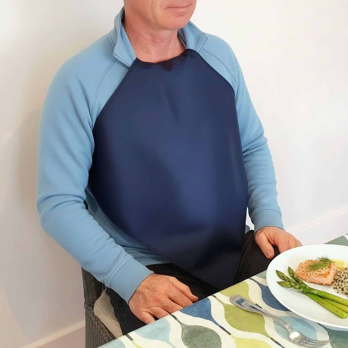 Man wearing a Care Designs navy waterproof napkin  over a light blue sweatshirt at a dining table with a colorful placemat.