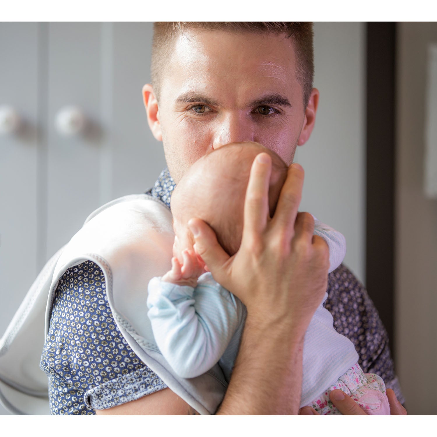 man with brown hair wearing a grey shirt holding a baby in a light blue top and flowery leggings on his shoulder on a Bibetta white and grey burp cloth